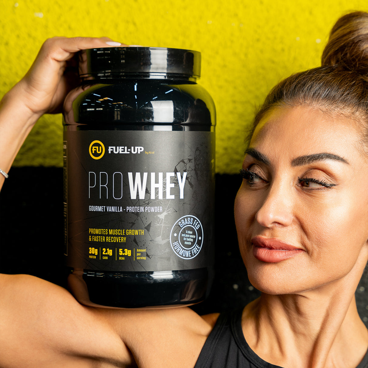 Woman holding a tub of vanilla whey protein powder on her shoulder in the gym